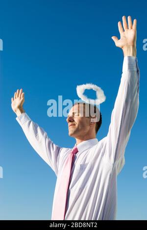 Engelhafte Geschäftsmann stehend Arme mit einem Heiligenschein schweben über seinem Kopf in den blauen Himmel angehoben Stockfoto