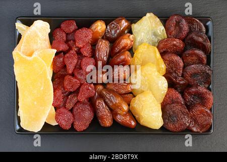 Getrocknete Stücke Mango-Obst; getrocknete Erdbeeren, Datteln, Pfirsiche und Aprikosen, die auf einer schwarzen Keramikplatte auf einer schwarzen Steinoberfläche liegen. Makro Stockfoto