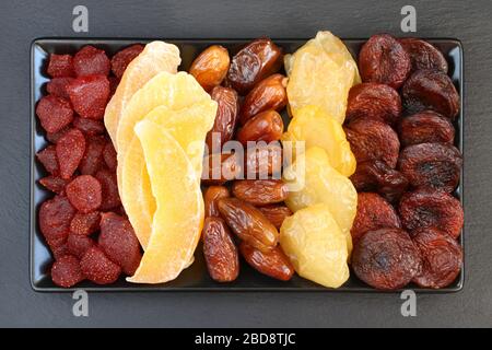 Getrocknete Stücke Mango-Obst; getrocknete Erdbeeren, Datteln, Pfirsiche und Aprikosen, die auf einer schwarzen Keramikplatte auf einer schwarzen Steinoberfläche liegen. Makro Stockfoto