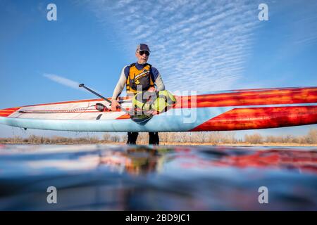 Senioren-Paddler in einem Wetsuit trägt ein Stand-Up-Paddleboard, nachdem er auf einem See in Colorado gepaddelt hat, Low-Angle-Action-Kameraansicht, rekre Stockfoto