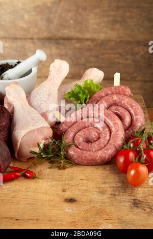 Unbehackte Wurstspulen und Hühnerbeine mit frischen Tomaten, Chilipapern und Gewürzen auf einem rustikalen Holzküchentisch mit Grillplatz Stockfoto