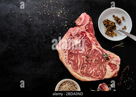 Roher marmorierter Knochen-in-Ribeye-Steak mit Gewürzen und Knoblauch in einer oben liegenden flachen Laienansicht mit Kopierraum auf dunklem Hintergrund Stockfoto