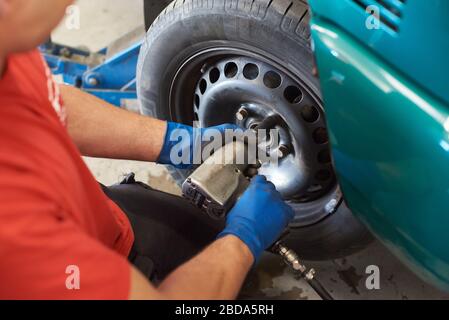 Nahaufnahme der männlichen Hände in blauen schmutzigen Handschuhen Installation Ansatzmuttern. Professioneller Mechaniker mit Schlagschrauber während der Arbeit in der Garage. Konzept des Autoservice. Stockfoto