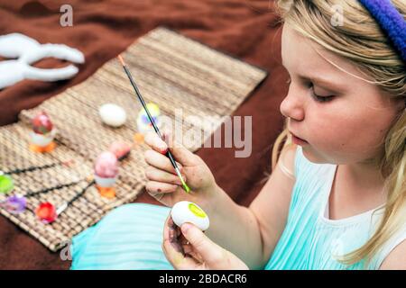 Niedliche kleine Mädchen, die am Ostertag auf der Decke auf Gras im Garten sitzen und auf den Eiern malen, tragen bunte Ohren. Stockfoto