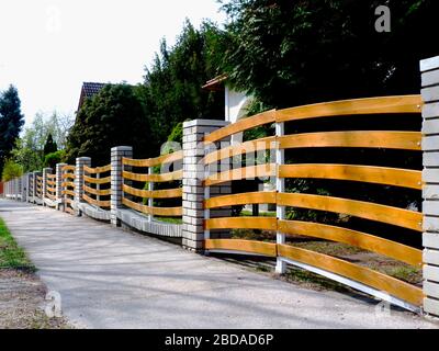 Pfostenzaun aus Backstein. Bunte gelbe Holzzaunverkleidungen. Üppige immergrüne Bäume im Hintergrund des Vorhofes. Sträucher und Kiefernbaum. Hausbesitz Stockfoto