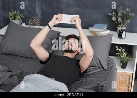Bärtiger Mann, der im Bett liegt und Tablette verwendet, während er zu Hause während der Quarantäne verbrachte Stockfoto