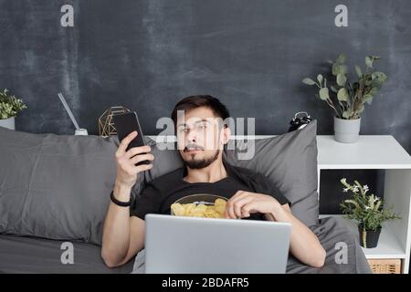 Langweiliger junger Student, der im Bett liegt und Chips isst, während er im Internet auf dem Smartphone surft, verschwendet er zu Hause Zeit während der Quarantäne Stockfoto