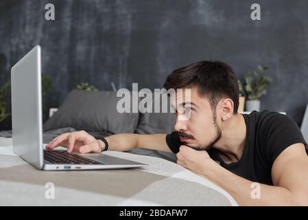 Langweiliger junger bärtiger Mann, der auf dem Laptop-Bildschirm schaut, während er auf Websites in Quarantäne surft Stockfoto