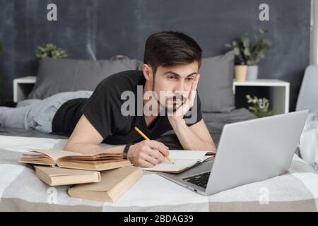 Langweiliger junger Mann, der im Bett liegt und Notizen im Arbeitsbuch macht, während er online mit einem Laptop zu Hause Themen studiert Stockfoto