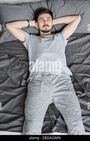 Penibler junger bärtiger Mann im Homewear, der mit den Händen hinter dem Kopf auf dem Bett liegt, während er sich bei der Quarantäne entspannt Stockfoto