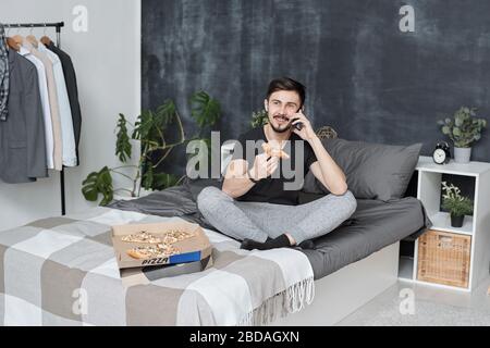 Fröhlicher, gutaussehender junger bärtiger Mann, der auf dem Bett sitzt und telefoniert, während er zu Hause Pizza isst Stockfoto