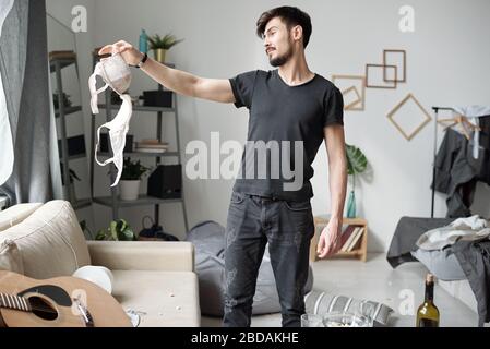 Der junge bärtige Mann in schwarz-legerer Kleidung hat nach einer Grobparty Brassiere beim Reinigen des Raumes gefunden Stockfoto