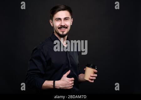 Porträt des fröhlichen, gutaussehenden jungen bärtigen Unternehmers mit Kaffeetasse, die vor schwarzem Hintergrund aufgebaggt ist Stockfoto