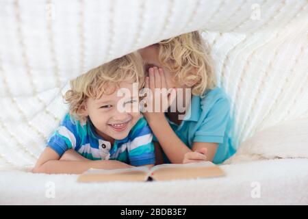 Lesebuch für Kinder im Bett unter der Strickdecke. Zwei Brüder spielen zusammen. Kinder gemütliches Zimmer im Hyggge-Stil. Kleiner Junge, der Hausaufgaben macht, bevor er schläft. Stockfoto