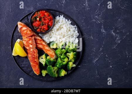 Meeresfrüchtemahlzeit: Gegrilltes Lachsfilet mit gekochtem Brokkoli und Reis mit Tomaten- und Spinatmischung, Zitronenkeile auf schwarzem Teller auf einer dunklen Zange Stockfoto