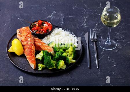 Platte aus gegrilltem Lachssteak mit gedämpftem Brokkoli, Langkornreis, Tomaten- und Spinatsoße, Zitronenkeile auf einem dunklen Betontisch mit einem Glas Stockfoto