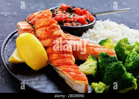 Nahaufnahme von frisch geröstetem Lachssteak mit Gemüse: Brokkoli-, Tomaten- und Spinatsoße und Langkornreis, Zitronenkeile auf einer schwarzen Platte Stockfoto