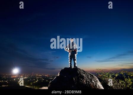 Tapferer Astronaut auf dem felsigen Hügel mit schönen blauen Himmel, Stadt Lichter auf dem Hintergrund. Schöner Spaceman in weißen Raum Anzug hält Helm und Blick auf Kamera. Konzept Raumreise. Stockfoto