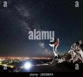 Kosmonauten stehen auf felsigen Hügel unter schönen Nachthimmel mit Sternen und Milchstraße. Space Traveler in Raum Anzug mit Helm und Blick auf die Nacht Stadtlichter. Konzept der Kosmonauten und der Nacht Stockfoto