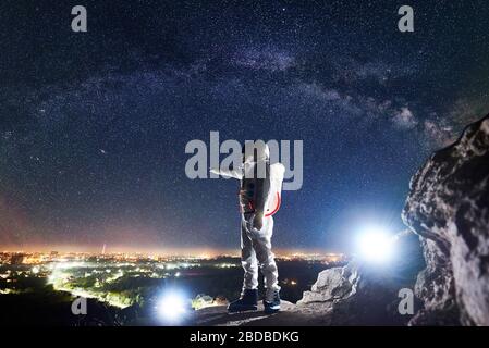 Männlicher Spaceman, der mit dem Finger auf die Stadt in der Nacht zeigt, während er auf felsigen Bergen unter dem fantastischen Nachthimmel mit Sternen und Milchstraße steht. Konzept der Kosmonauten und Raumfahrt. Stockfoto