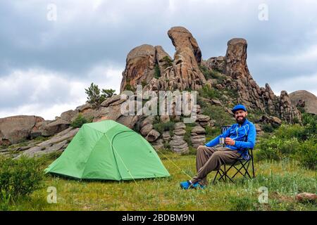 Ein fröhlicher Tourist auf einem Zeltplatz sitzt auf einem Klappstuhl in der Nähe eines grünen Zeltes mit einem Metallbecher in seinen Händen. Camping mit Blick auf ungewöhnliche, schöne Felsen. T Stockfoto