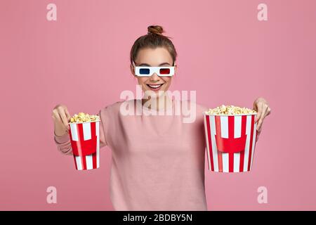 Junge schöne kaukasische Frau trägt rot-blaue 3d-Brille und hält zwei Eimer Popcorn isoliert auf rosa Hintergrund. Mädchen nahm Popcorn für sich und für einen Freund Stockfoto