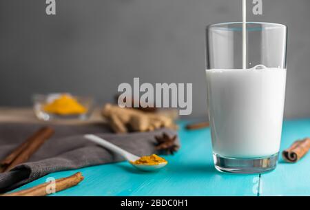 Milch wird in ein Glas für goldene Milch, Milch mit Kurkuma, Gewürze, mit Trinkrohr, auf blauem Holzhintergrund, mit Kopierraum gegossen Stockfoto