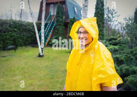 Porträt der schönen jungen erwachsenen kaukasusfrau unter hellgelbem Regenmantel und Schweifdecken, die während des kalten Frühlings-Regentags zu Hause im Garten spazieren Stockfoto