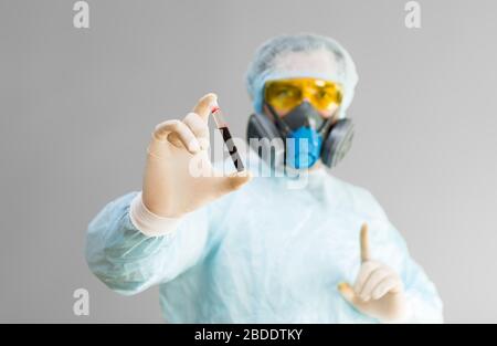 Foto eines Arztes in Schutzkleidung hält ein Reagenzglas mit einer Probe infizierten Blutes in der Hand und zeigt ein Aufmerksamkeitszeichen mit dem Finger. Healthcar Stockfoto