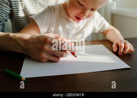 Vater, der dem Kind beigebracht hat, mit den Händen zu schreiben oder zu zeichnen. Roter Bleistift und Papier auf Holztisch. Homeschooling Stockfoto