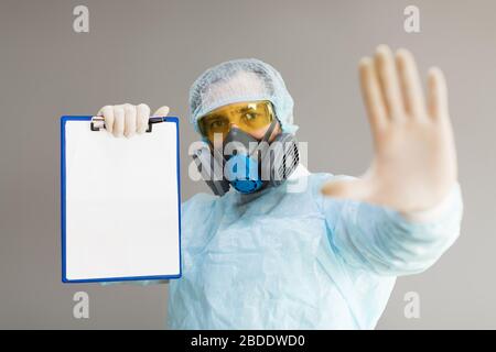 Ein Arzt in Atemschutzmaske und Schutzkleidung zeigt ein Stoppschild mit seiner Handfläche und hält ein Papier mit einer Diagnose. Mock-up. Stockfoto