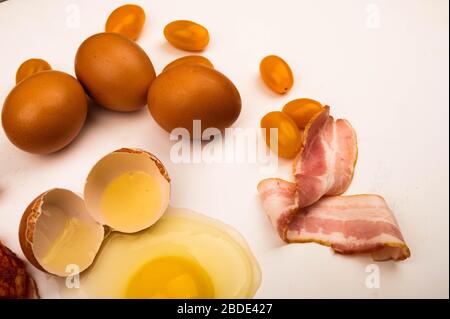 Zerbrochenes Hühnerei, Hühnereier, Speckscheiben und Tomaten auf weißem Hintergrund. Nahaufnahme Stockfoto