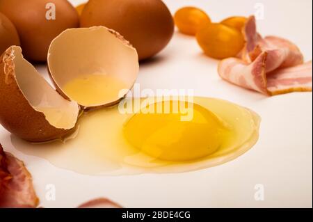 Zerbrochenes Hühnerei, Hühnereier, Speckscheiben und Tomaten auf weißem Hintergrund. Nahaufnahme Stockfoto