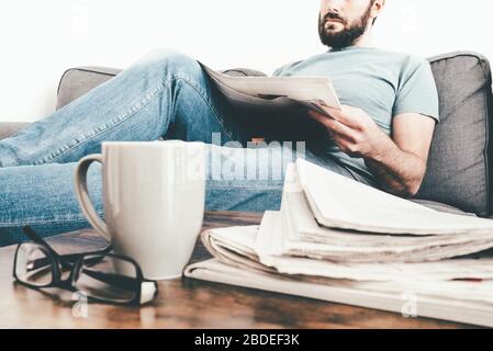 Man entspannt sich auf dem Sofa und liest eine Zeitung mit einem Stapel Zeitungen und einem Kaffeebecher im Vordergrund Stockfoto