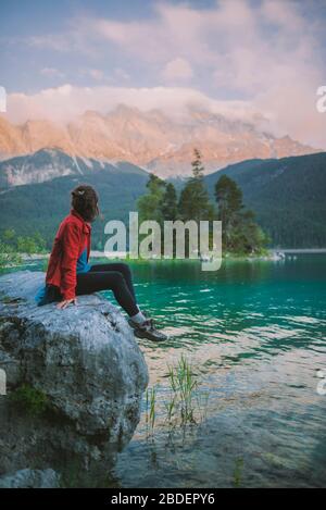 Deutschland, Bayern, Eibsee, Junge Frau auf Felsen sitzend und Blick auf den malerischen byÂ EibseeÂ See in den bayerischen Alpen Stockfoto