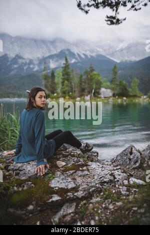 Deutschland, Bayern, Eibsee, Portrait einer jungen Frau, die am Ufer des Eibsees in den bayerischen Alpen auf Felsen sitzt Stockfoto