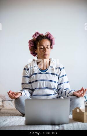 Frau mit Lockenwicklern meditiert auf dem Bett vor Notebook Stockfoto