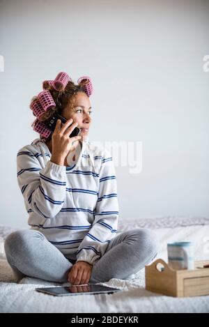 Frau mit hairÂ curlersÂ sitzt auf dem Bett und spricht auf dem Handy Stockfoto