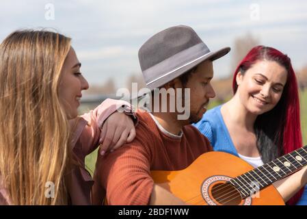 Ein hispanischer Junge in einem Hut spielt neben zwei kaukasischen Mädchen in einem Stadtpark die Gitarre Stockfoto