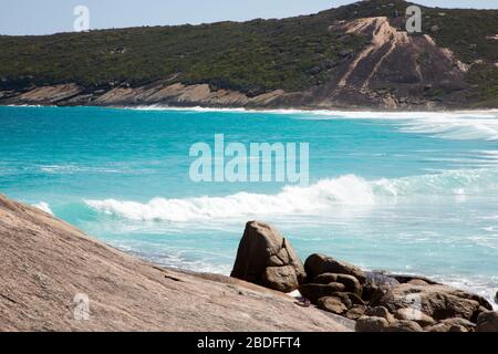 Blaue Wellen, grüne Hügel und wilde Felsen und Steine der Hellfire Bay im Cape Le Grand National Park in Western Australia; getragene Turnschuhe im Vordergrund Stockfoto