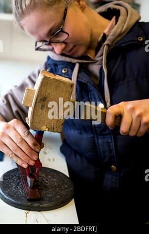 In der Werkstatt stehender Sattler mit Handwerkzeug und Holzhammer, der Löcher in den Lederriemen stanzt. Stockfoto