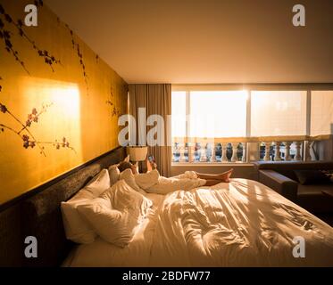 Erwachsene Frauen, die in der Sonne am frühen Morgen auf dem Hotelbett liegen und auf ein Smartphone blicken Stockfoto