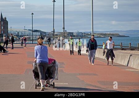 Portobello Beach, Edinburgh, Schottland, Großbritannien. April 2020. 18 Grad und voller Sonnenschein brachten Sonnenanbeter ans Meer, die meisten sozialen Distanzierungen, aber nicht die Menschenmassen, die erwartet würden, wenn es nicht den Coronavirus Lockdown gäbe und die Menschen hauptsächlich spazieren oder sitzen, anstatt auf dem Sand zu liegen. Ein männlicher und weiblicher Polizist auf der Promenade für einen Routinepassgang. Credit: Arch White/Alamy Live News Stockfoto