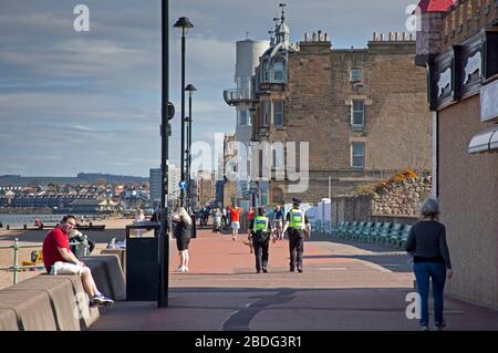 Portobello Beach, Edinburgh, Schottland, Großbritannien. April 2020. 18 Grad und voller Sonnenschein brachten Sonnenanbeter ans Meer, die meisten sozialen Distanzierungen, aber nicht die Menschenmassen, die erwartet würden, wenn es nicht den Coronavirus Lockdown gäbe und die Menschen hauptsächlich spazieren oder sitzen, anstatt auf dem Sand zu liegen. Ein männlicher und weiblicher Polizist auf der Promenade für einen Routinepassgang. Credit: Arch White/Alamy Live News Stockfoto