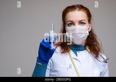 Stock Foto Porträt eines rothaarigen professionellen Wissenschaftlers in Schutzmaske, weißem Mantel und Handschuh, der eine Spritze hält, die mit Impfstoff gegen gefüllt ist Stockfoto