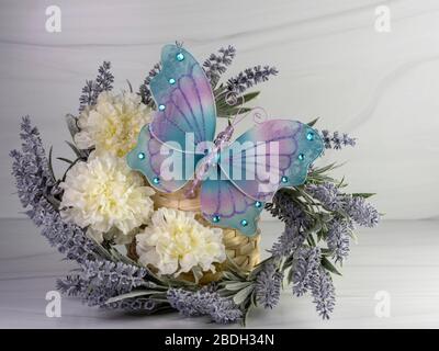 Violetter Lavendelkranz mit weißen Nelken und einem großen blauen und violetten Schmetterling aus Mesh, der mit Edelsteinen verziert ist, die auf einem sandsteinfarbenen Hintergrund sitzen. P Stockfoto