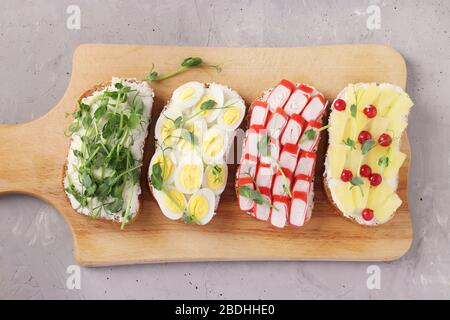 Vier Sandwiches auf Toast mit Erbsen, Mikrogrüns, Ananas, rotem Currant, Krabbenstäbchen und Wachteleiern auf Holzbrett auf grauem Betonhintergrund Stockfoto