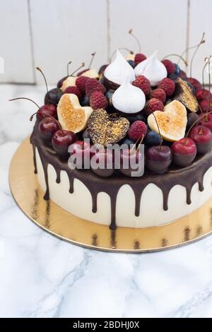 Vanillekäsekuchen mit geschmolzener dunkler Schokolade, frischen Beeren, Kirschen, Schokoladenherzen und Baiser. Weißer rustikaler Hintergrund. Stockfoto