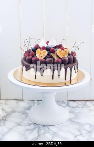 Vanillekäsekuchen mit geschmolzener dunkler Schokolade, frischen Beeren, Kirschen, Schokoladenherzen und Baiser. Weißer rustikaler Hintergrund. Stockfoto