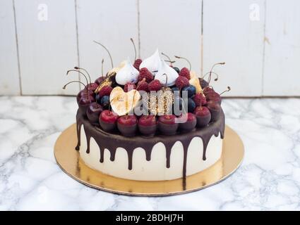 Vanillekäsekuchen mit geschmolzener dunkler Schokolade, frischen Beeren, Kirschen, Schokoladenherzen und Baiser. Weißer rustikaler Hintergrund. Stockfoto